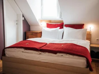 A neatly made bed with white sheets and red blankets in a cozy bedroom with a slanted ceiling and a window above the headboard. Two small nightlights are on each side.