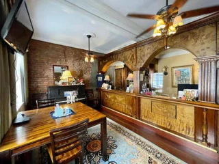 A room with wooden furniture, a table with chairs, a ornate reception counter, brick and decorated walls, and a ceiling fan. There are various items on the counter and a TV mounted on the wall.