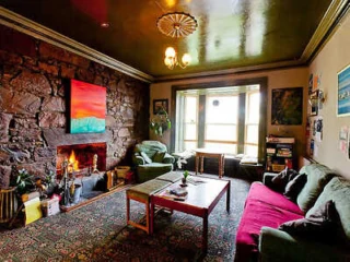 A cozy living room with a lit fireplace, stone wall, colorful artwork, a green armchair, a red couch, and a wooden coffee table on a patterned rug. The room has a bay window illuminated by natural light.