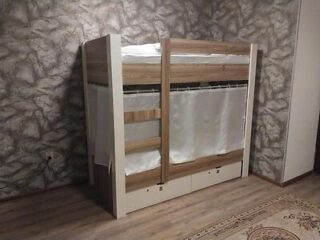 A wooden bunk bed with a top bed rail and bottom bed curtains in a room with stone-patterned wallpaper and a hardwood floor. There are drawers under the bottom bed.