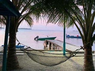 A hammock is strung between two palm trees, facing a small wooden pier that extends into calm waters. A small motorboat is docked at the pier, and the sky is tinged with pink and orange hues.