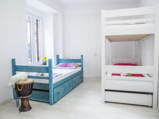 A minimalistic bedroom with a twin bed, a bunk bed, a djembe drum with a plush toy on it, and a small flower pot on a bedside table. The room features white walls and light wood flooring.