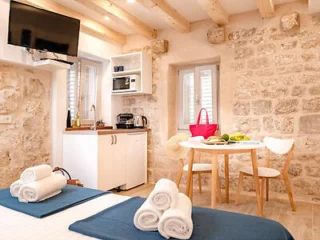 A cozy room with a stone wall interior. The room features a bed with folded towels, a small dining area with a table and chairs, a kitchen corner with a microwave and coffee maker, and a mounted TV.