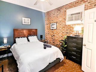 A cozy bedroom features a double bed with white bedding, a dark wooden headboard, blue-painted and brick walls, a dresser, a small window unit air conditioner, and a wooden nightstand with a lamp.
