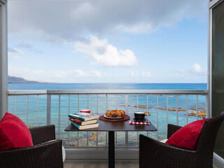 A small table with books, pastries, and a coffee set, positioned between two wicker chairs with red cushions, overlooks a serene ocean view through a glass railing.