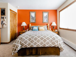 A tidy bedroom with an orange accent wall, a bed with patterned bedding, two nightstands with lamps, and framed pictures above the bed. The room has a window on the right and a closed door on the left.