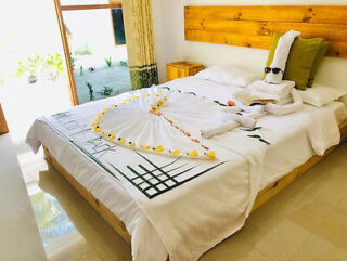 A neatly made bed with white linens, decorated with a towel sculpture wearing sunglasses, and surrounded by small yellow flowers. A wooden headboard and a patio door to the left are visible.