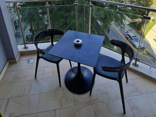 A small balcony with a black round table and two matching chairs, offering a view of the surrounding greenery and street below