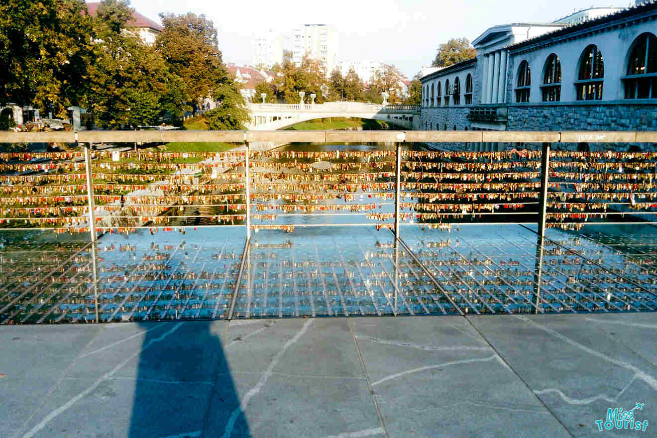 Bridge with numerous padlocks attached to railings, trees and buildings in the background, and a clear blue sky overhead.