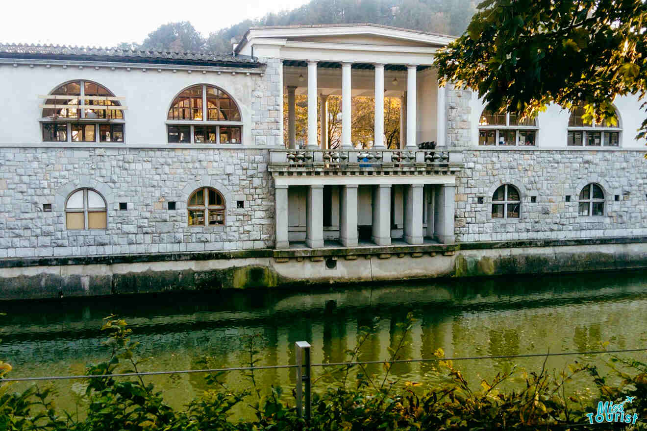 A large stone building with tall columns sits along a calm green river, framed by trees in the foreground.