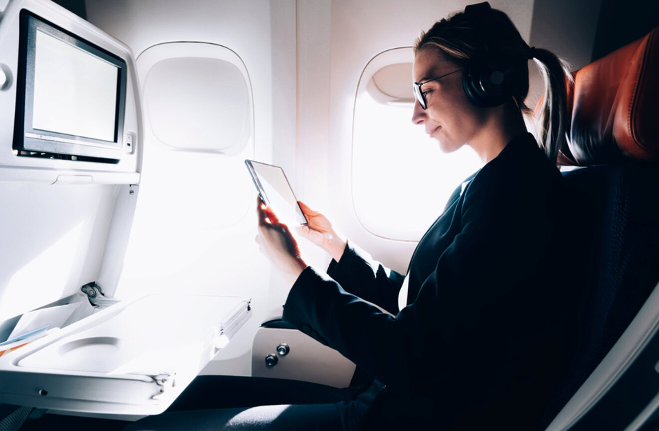 A woman wearing headphones and glasses reads on a tablet in an airplane seat with a fold-down tray and an in-flight entertainment screen beside her.