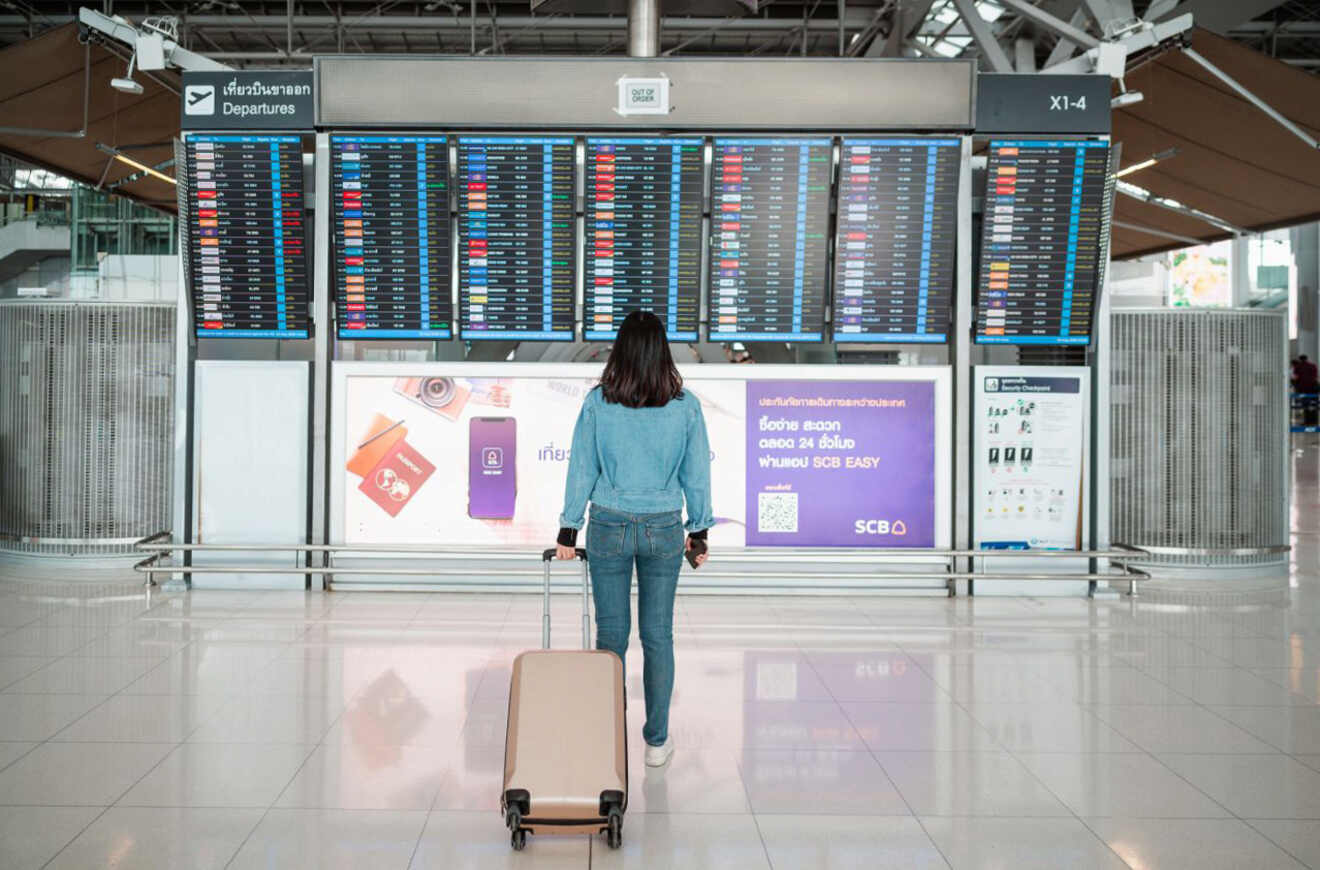 Person standing in front of a large departure board at an airport, holding a suitcase.