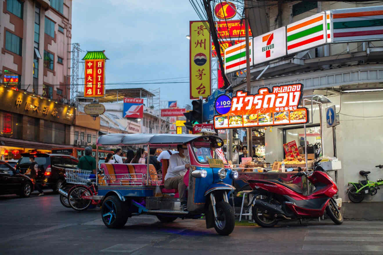 A busy outdoor street market at dusk with a tuk-tuk parked near food stalls, neon signs, and various storefronts.