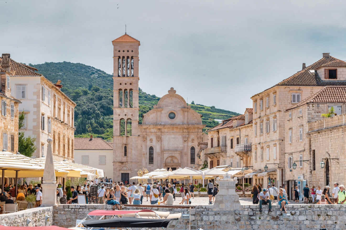 A bustling town square features historic buildings, a central clock tower, outdoor seating, and people enjoying the area, set against a backdrop of green hills.