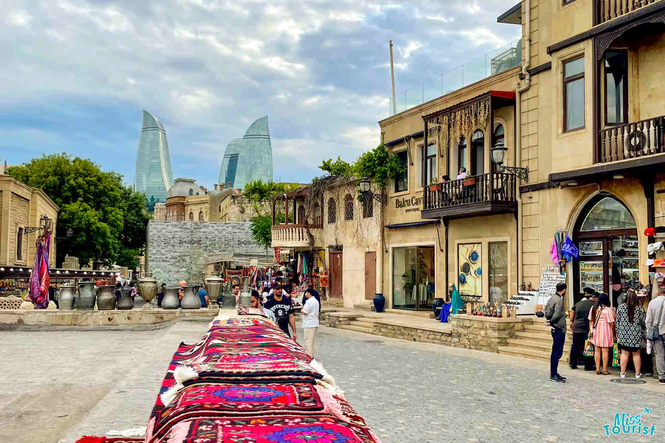 A street scene in Baku, Azerbaijan, featuring vendors with traditional carpets, historic buildings, and the Flame Towers visible in the background.
