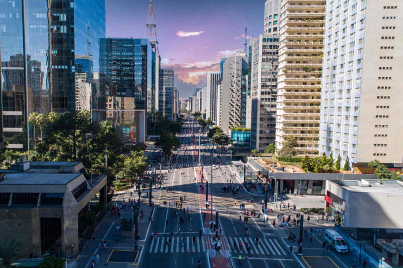 Aerial view of a busy city street flanked by modern high-rise buildings with pedestrians and vehicles visible, under a partly cloudy sunset sky.