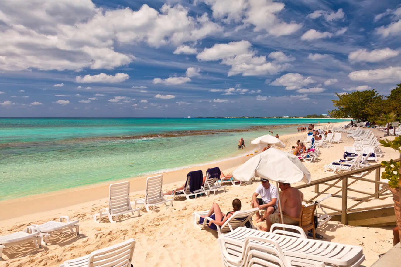 A sandy beach with lounge chairs and umbrellas lined up, people relaxing under umbrellas and swimming in the clear turquoise water. The sky is partly cloudy.