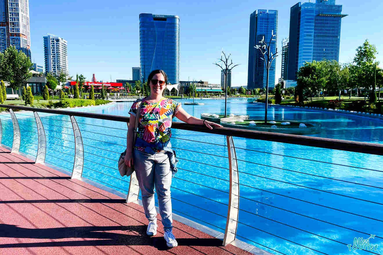 author of the post standing on a bridge over a blue body of water, with modern high-rise buildings and greenery in the background under a clear sky.