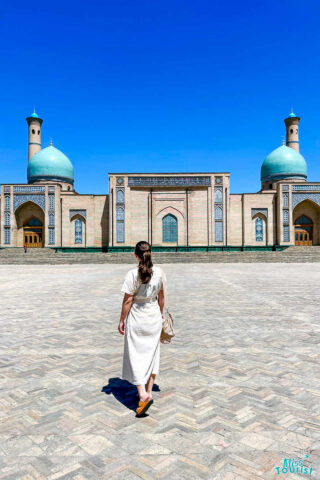 author of the post in a white dress walks towards a historic building with two turquoise-domed towers under a clear blue sky.