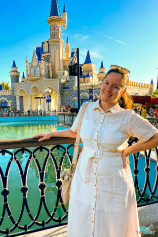 author of the post in a white dress stands by a decorative railing in front of a castle-themed structure on a sunny day.