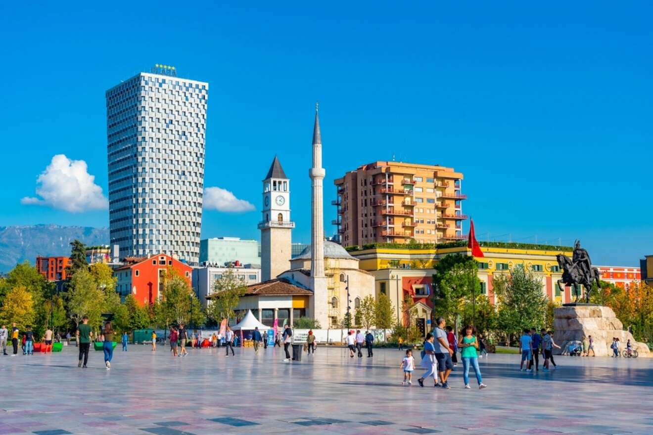 A diverse group of people walk across a spacious square with a statue, surrounded by colorful buildings, a mosque, and a clock tower under a clear blue sky.