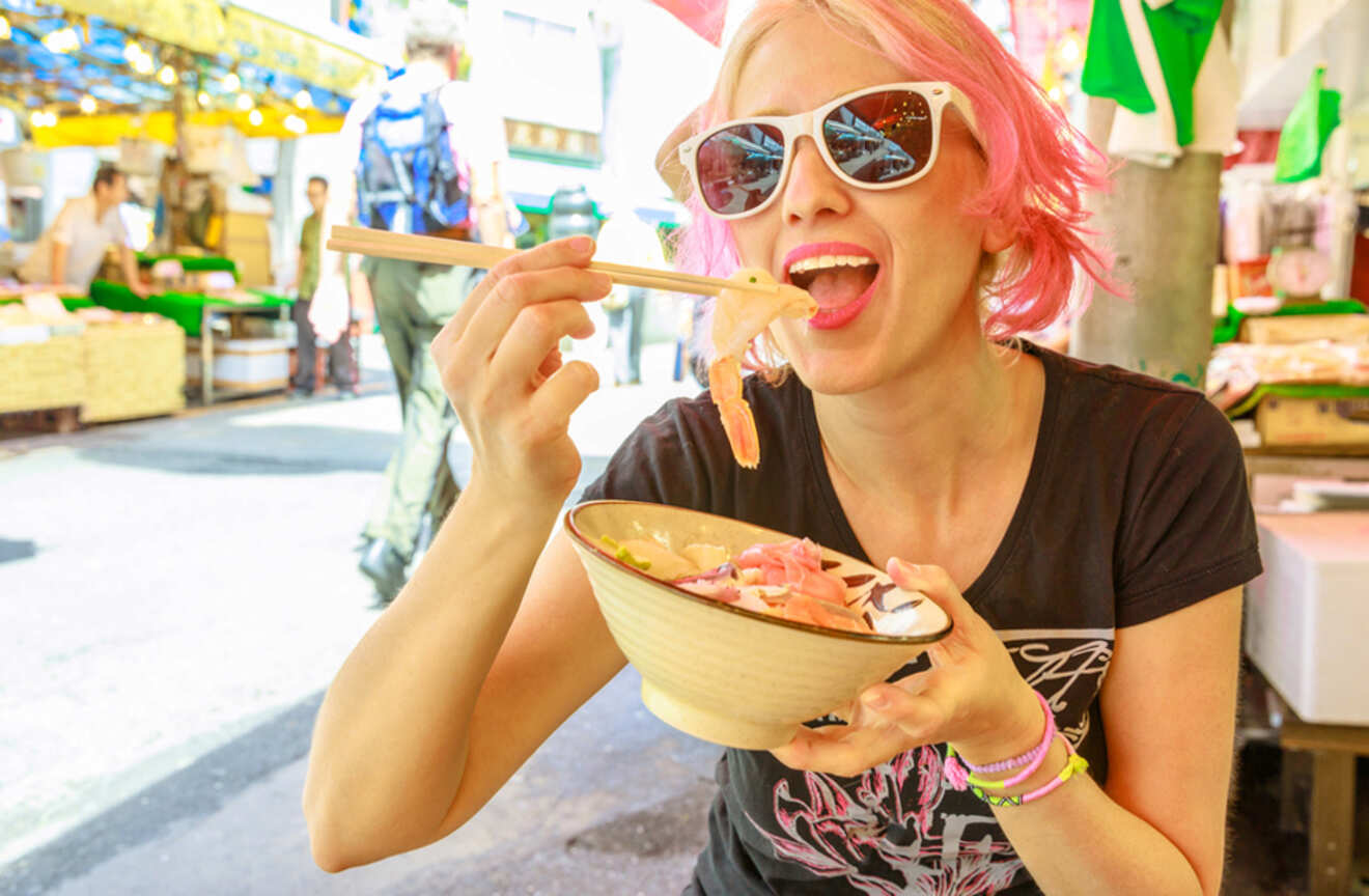 Woman with pink hair and sunglasses enjoying a bowl of seafood at a street market in Tokyo, Japan.