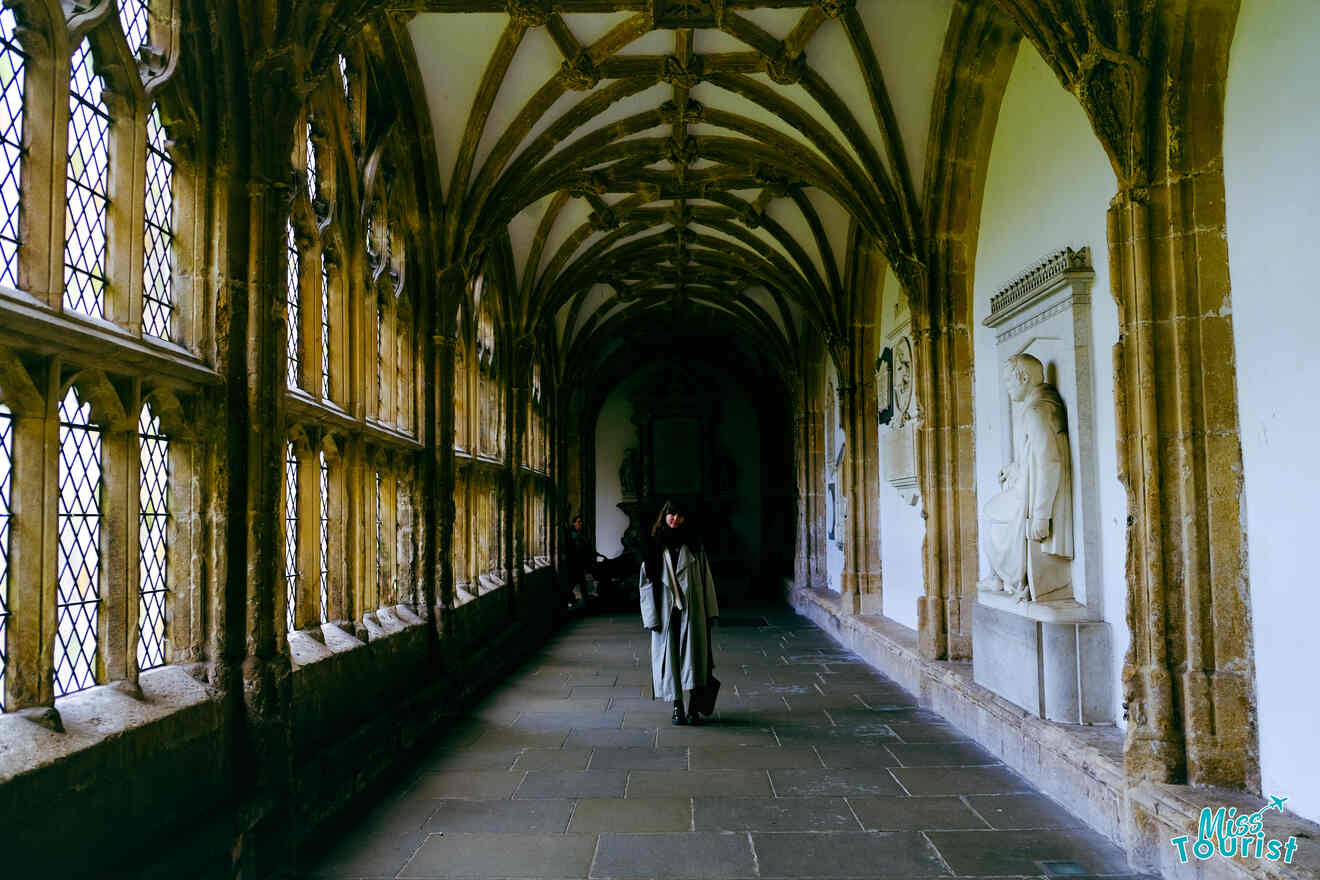 The writer of the post walking through a stone corridor with arched windows and historical sculptures at Wells Cathedral