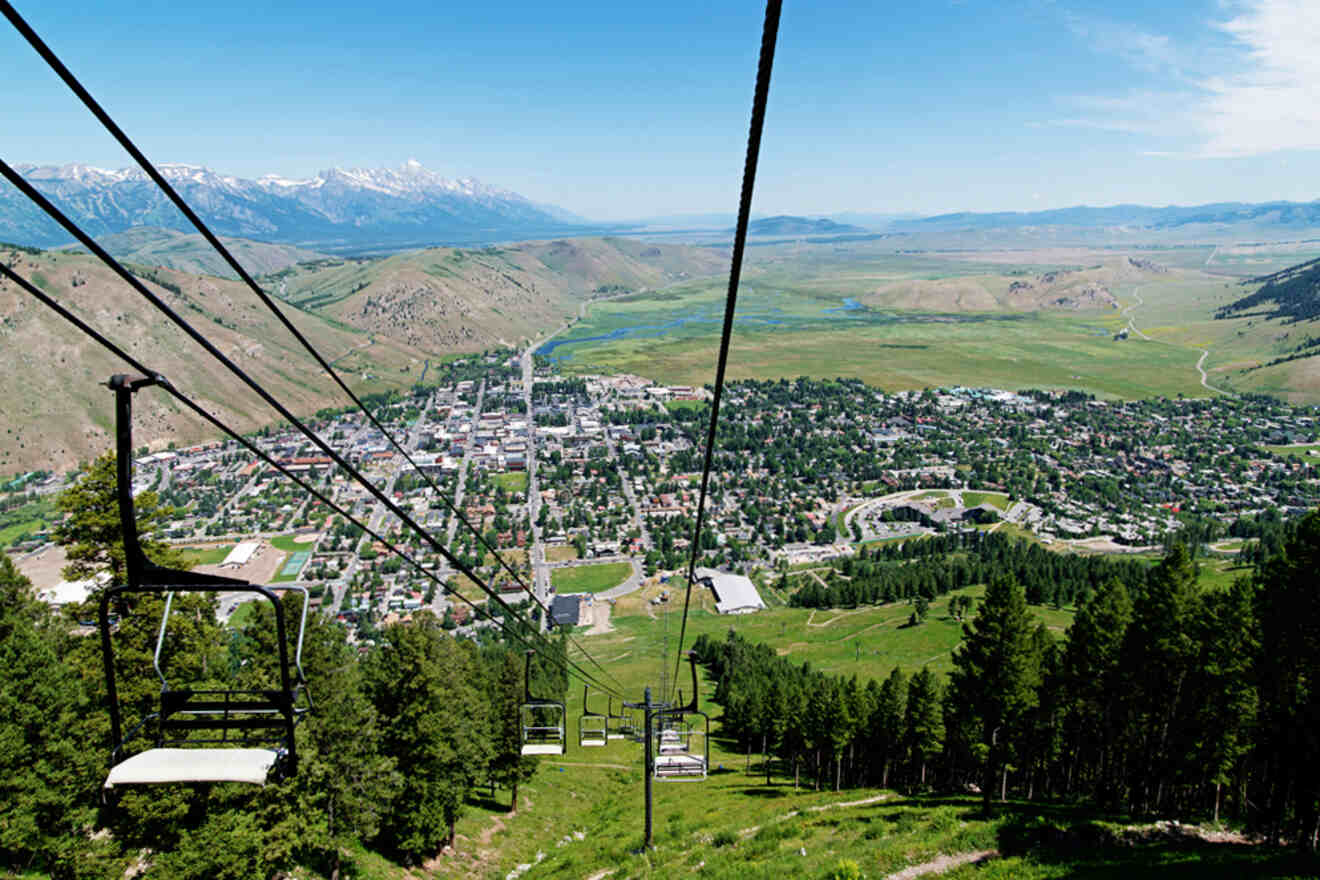 Scenic view from a ski lift overlooking a town with buildings, greenery, and mountains in the background under a clear blue sky.