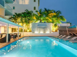 An outdoor swimming pool area with loungers, a waterfall feature, and palm trees, adjacent to a building at dusk.