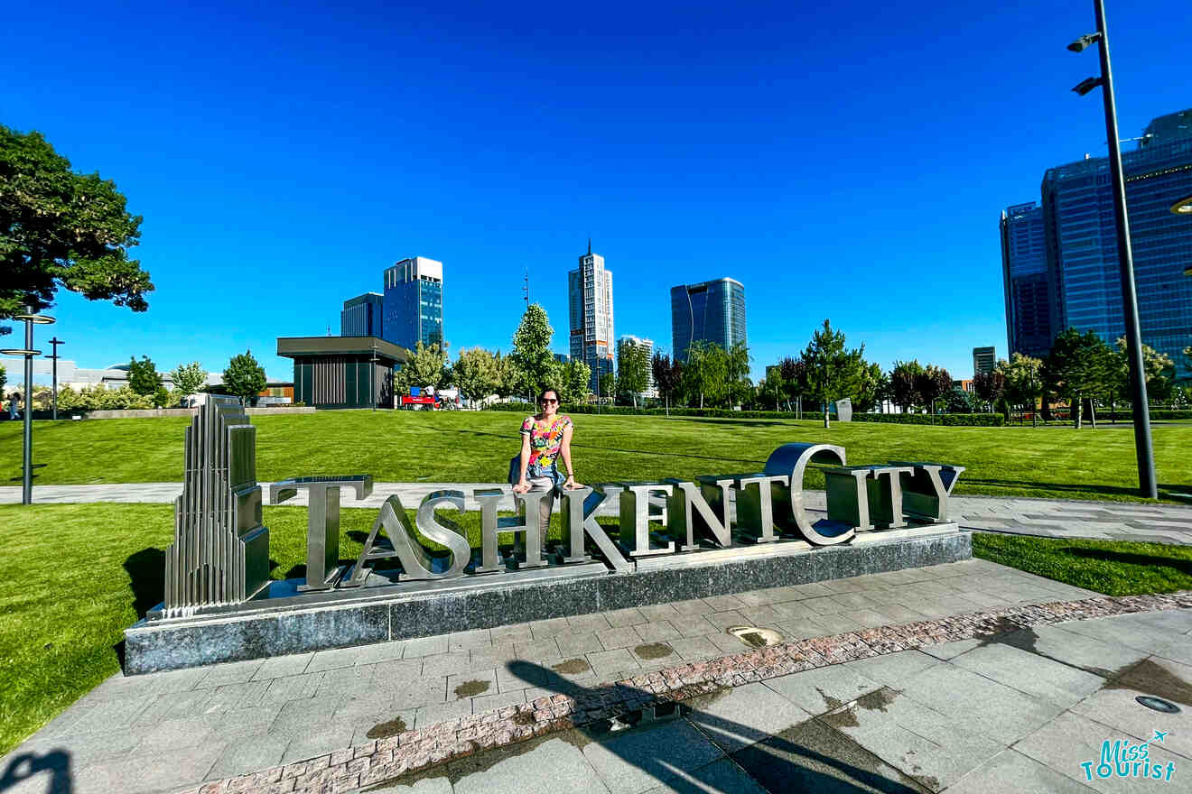 author of the post sits on a large "TASHKENT CITY" sign in a park, with modern buildings and a clear blue sky in the background.