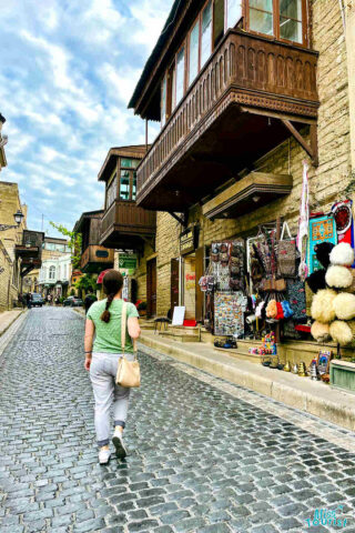author of the post with a green shirt and white pants walks on a cobblestone street lined with buildings and various outdoor market stalls on the right side.