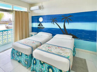 Two single beds with decorative covers in a bright room featuring a wall mural of a beach scene. The room has a white tile floor, beige curtains, and a view of a balcony outside the window.