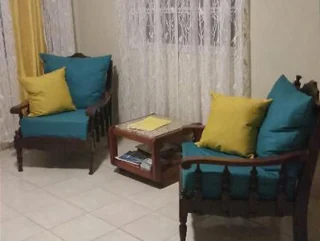 Two wooden armchairs with blue cushions and yellow throw pillows are placed next to a small wooden table. The table has books and a yellow item on top. Sheer curtains cover the window behind.