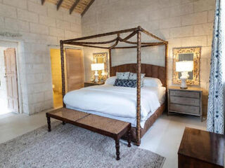 Um quarto com cama de dossel, roupa de cama branca, duas mesas de cabeceira com abajures, um banco aos pés da cama e paredes de pedra clara.