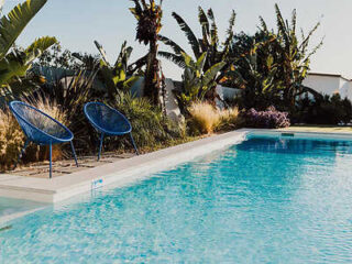 Outdoor swimming pool with two blue chairs on the poolside, surrounded by tropical plants and trees. The scene is sunny and tranquil.