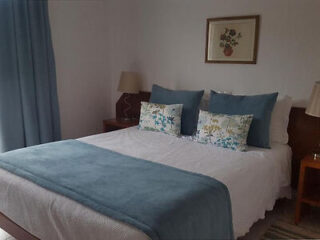 A neatly made bed with white sheets, blue pillows, and a blue throw blanket. Two bedside tables each have a lamp, and a floral picture hangs above the bed. Blue curtains are on the left.