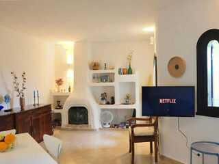 A minimalist living room with white walls, a wall-mounted TV displaying "Netflix," a rustic wooden sideboard, a modern fireplace, and built-in shelves. A table with fruit and a chair are also visible.