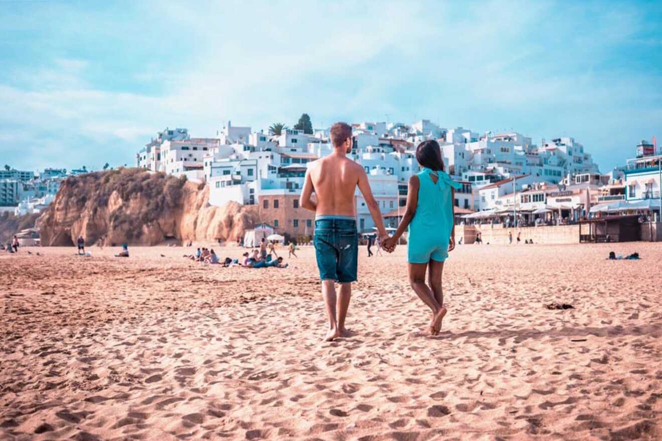 A couple holds hands while walking on a sandy beach with a coastal village of white buildings in the background.