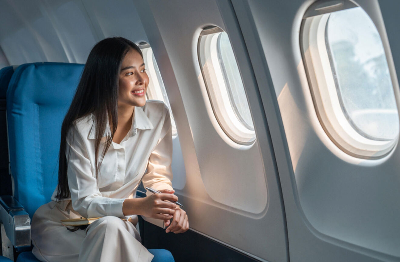 A woman with long hair sits comfortably by the airplane window, smiling and looking outside.