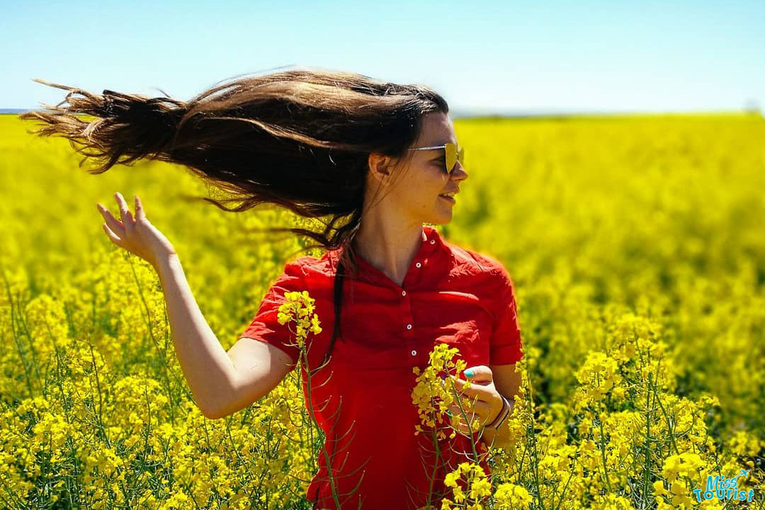 Yulia, la fundadora de este sitio web, con una camisa roja, se encuentra en un campo de flores amarillas, echándose el pelo largo hacia atrás y usando gafas de sol.