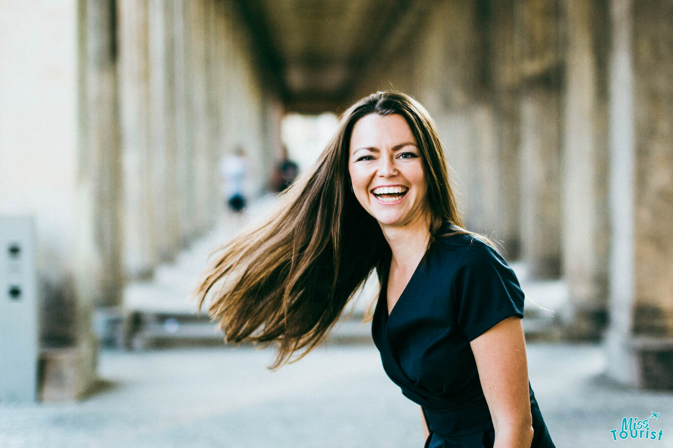 Yulia, la fundadora de este sitio web, con cabello largo y un vestido negro, sonríe y se inclina hacia adelante en un pasillo al aire libre con columnas.