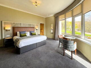 A spacious hotel room with a large bed, two chairs and a small round table by a wide window. The room is carpeted and has neutral-colored walls and bedding.