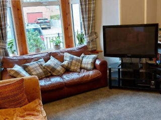 Living room with a brown leather sofa covered in plaid cushions, a flat-screen TV on a black stand, and large windows with checkered curtains.