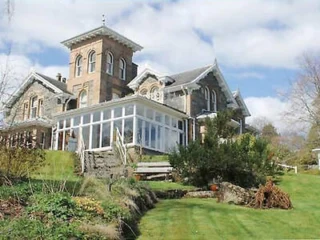 Large stone house with a tall tower and extensive glass-walled addition on a slight hill, surrounded by green lawn and landscaping under a partly cloudy sky.
