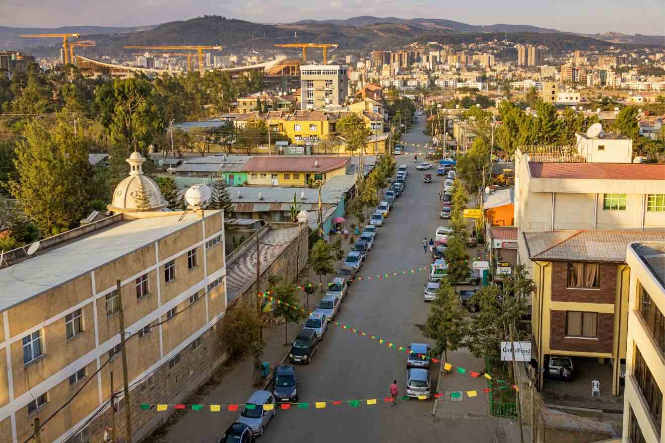 Aerial view of a street lined with cars and buildings in Addis Ababa, Ethiopia, with the cityscape and construction cranes visible in the background. Colorful flags are strung across the street.