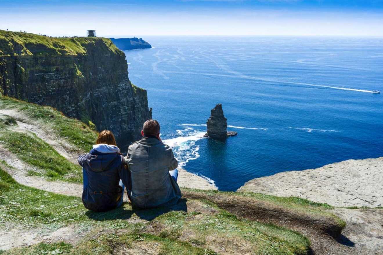 Two people sit on a grassy cliffside overlooking the ocean with a tall rock formation rising from the water. A distant boat sails near the horizon. The sky is clear and blue suggests a sunny day.