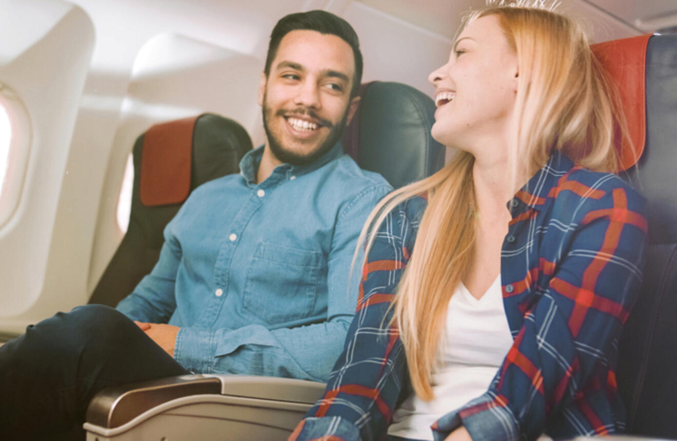 A man and a woman seated next to each other in an airplane are engaged in a lively conversation, both smiling.