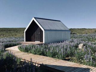 A small modern house with a slanted metal roof is situated in a field of purple flowers. A wooden path leads to the entrance.