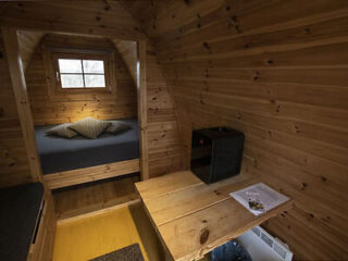 Interior of a small wooden cabin with a bed, a window, a table with papers on it, and a small storage unit. The space is furnished with basic necessities.