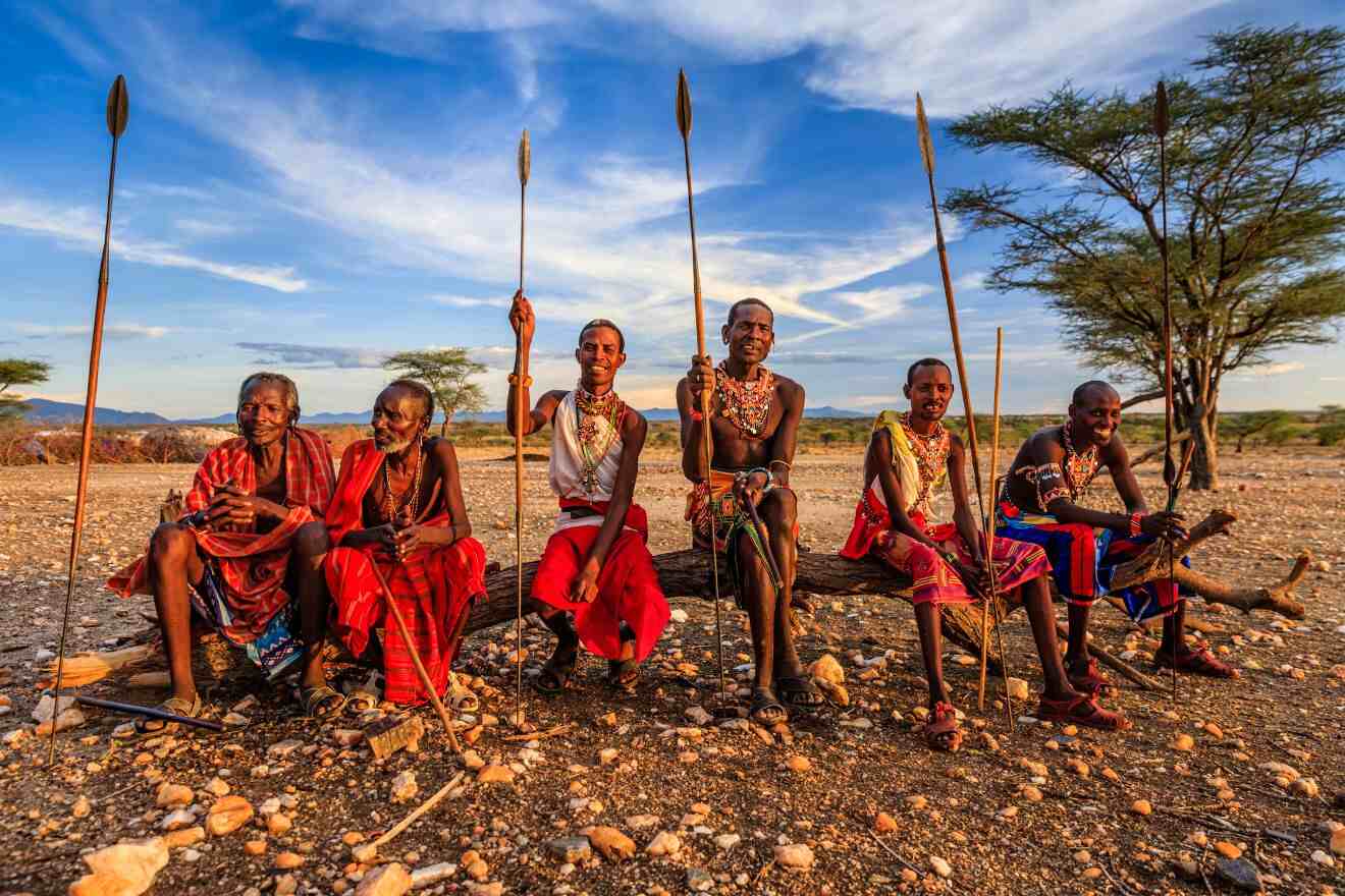 A group of people in traditional clothing sit outdoors on a log, holding spears, with a dry landscape and trees in the background.